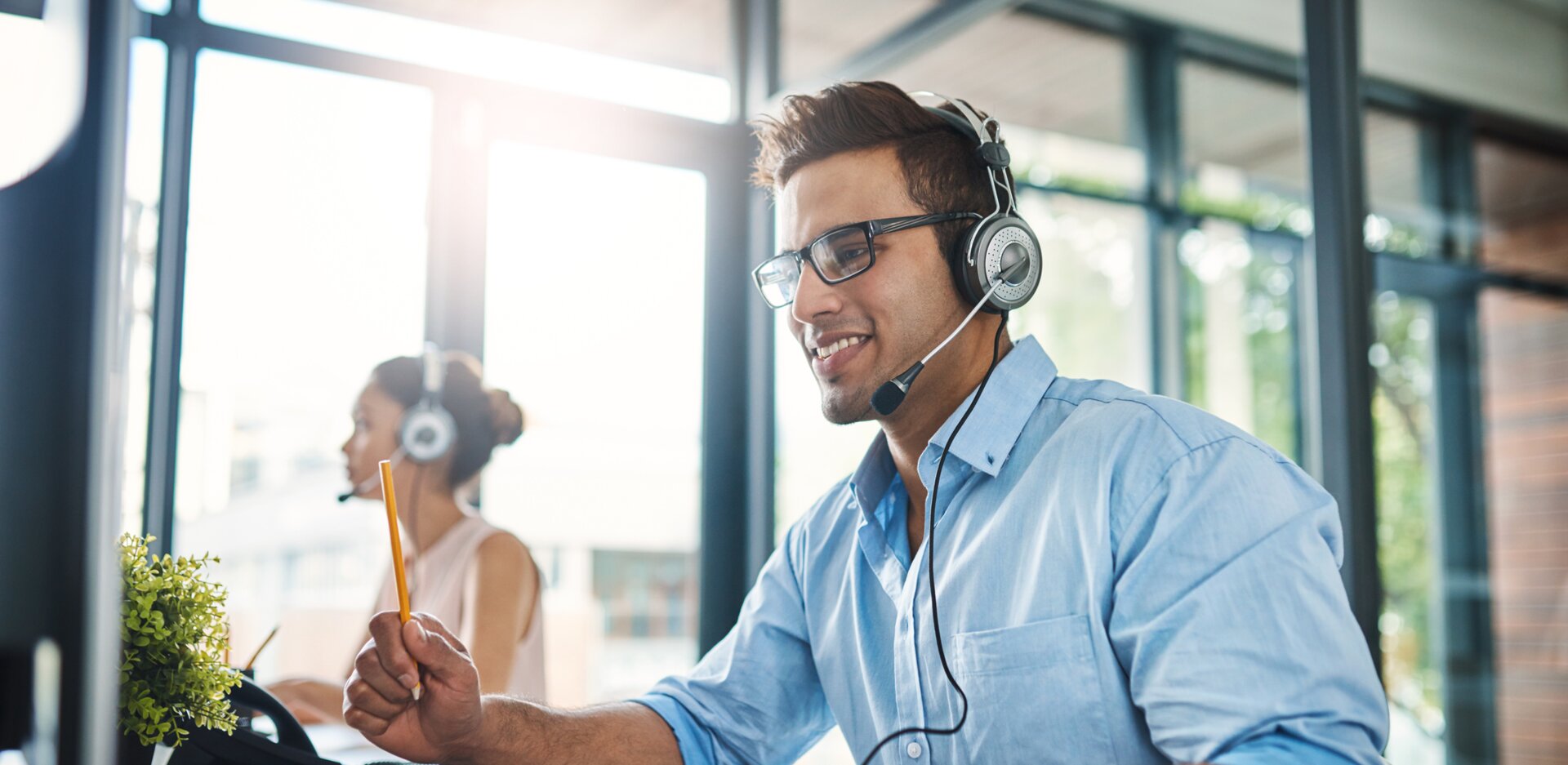 © iStock-1132874986_PeopleImages (Cropped shot of a handsome young man working in a call center with a female colleague in the background)